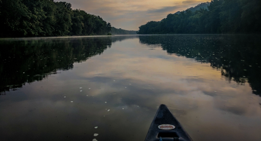 The very tip of a canoe is pictured in the foreground, floating on calm water, which reflects trees on the shore and a gray and yellow sky.